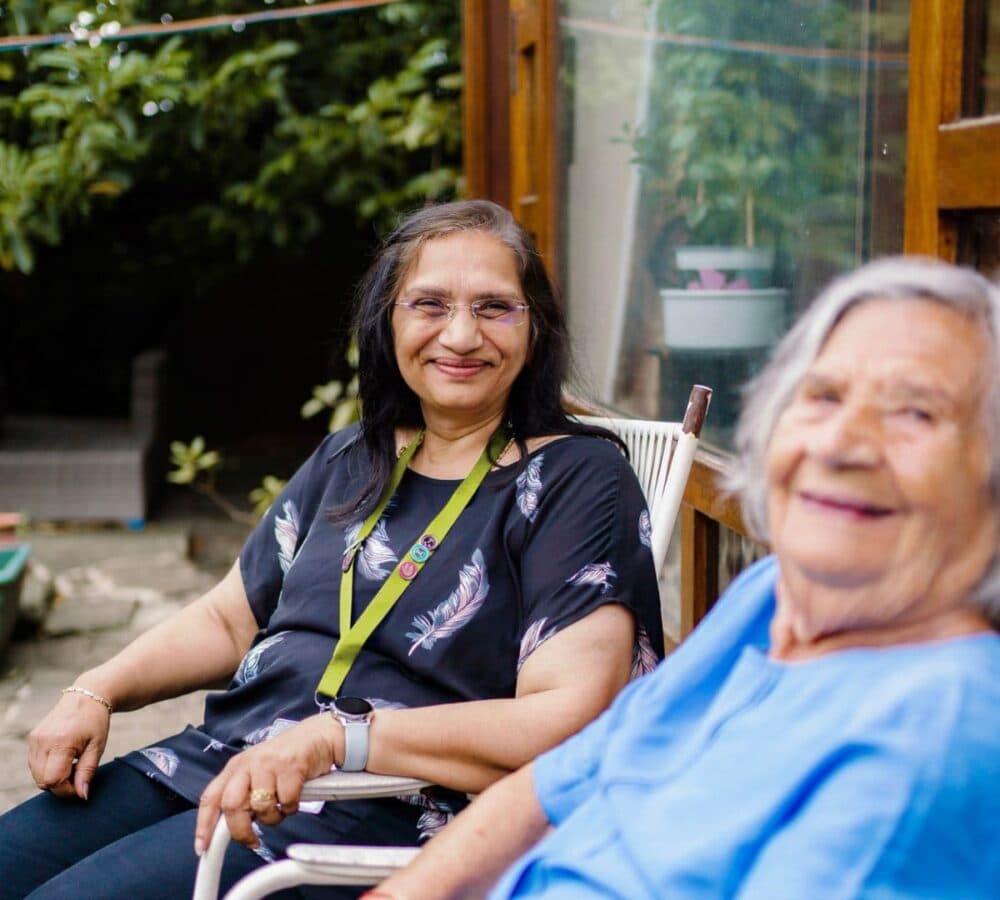 two women sitting on a chair outside the house and with plants at the back