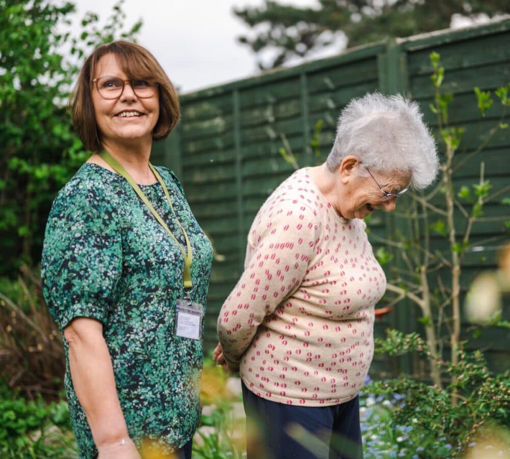 An older female adult with grey hair and eyeglasses walking in the garden with her younger female carer with short hair and wearing green top both happy and smiling