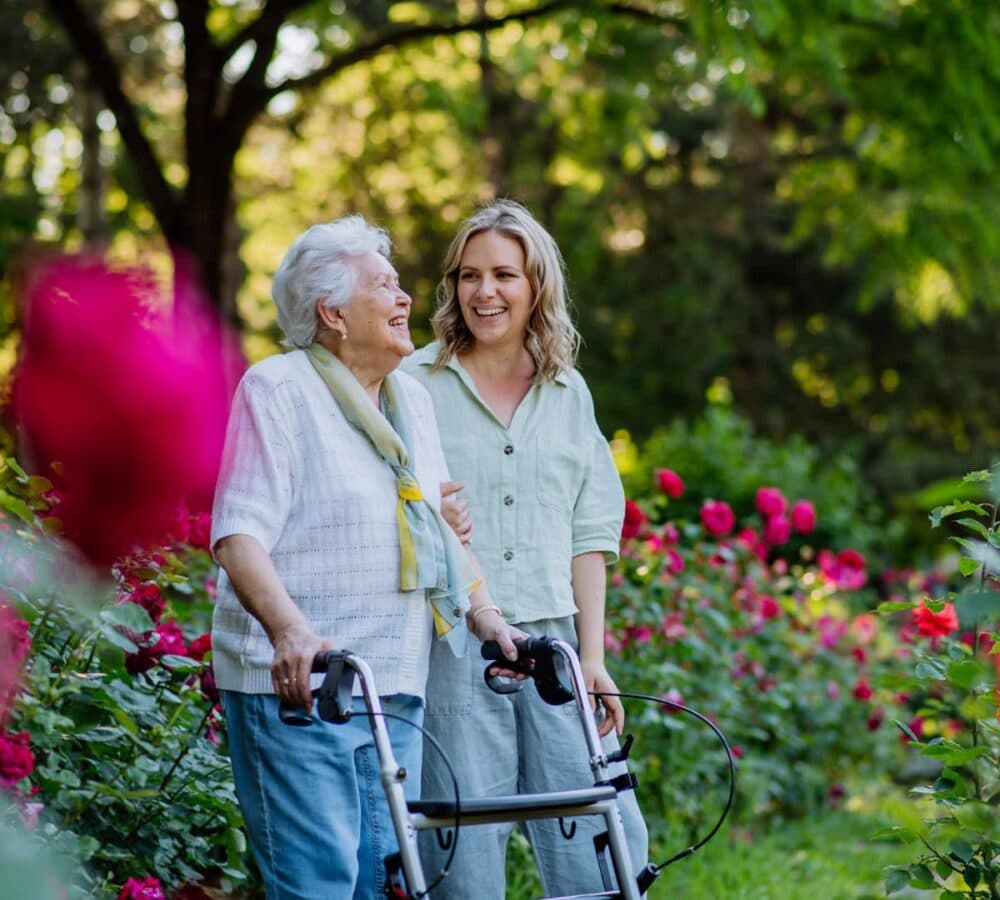TAn older female adult using a walker and with grey hair smiling while walking outside with her younger female carer happy and smiling while surrounded by greeneries and flowers outdoors