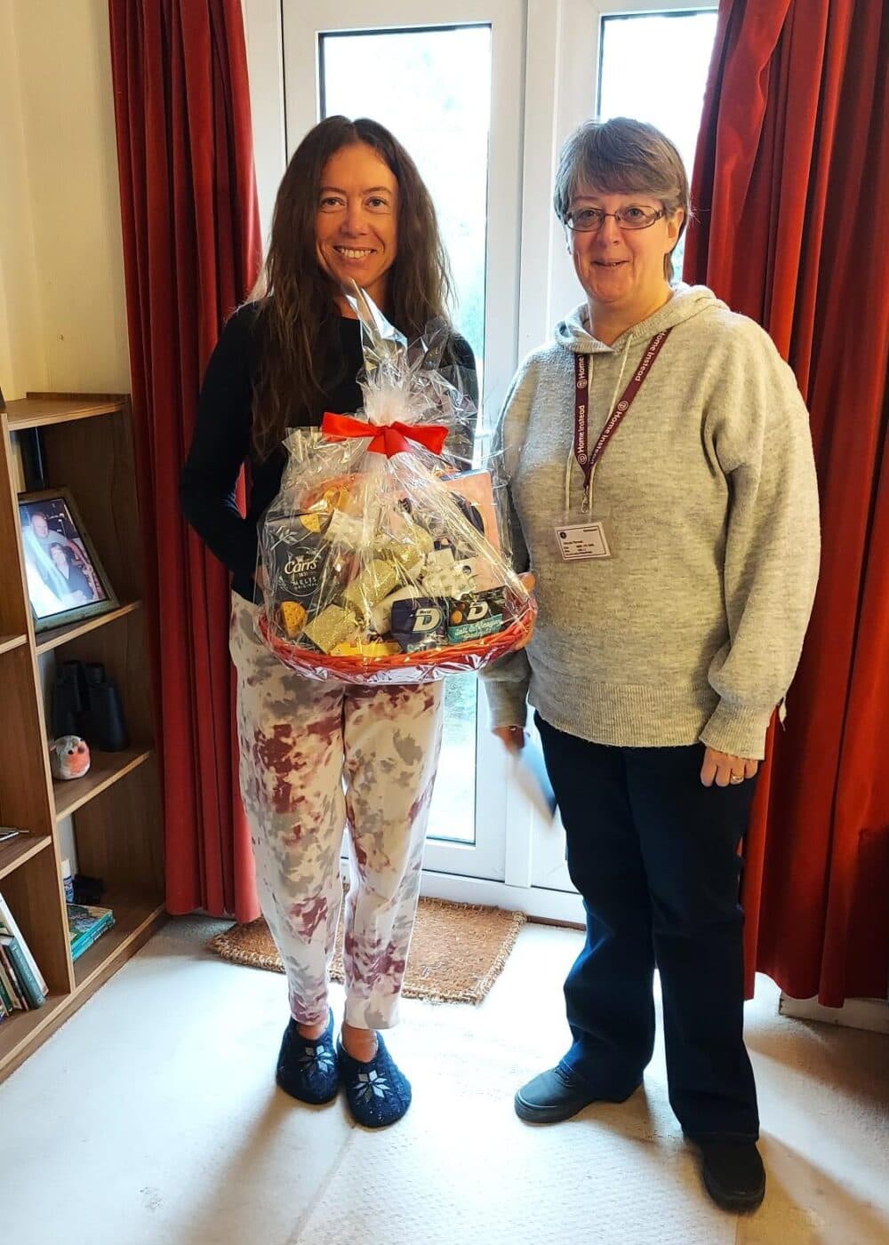 Two women stand indoors; one holds a gift basket wrapped in cellophane and tied with a red ribbon. - Home Instead