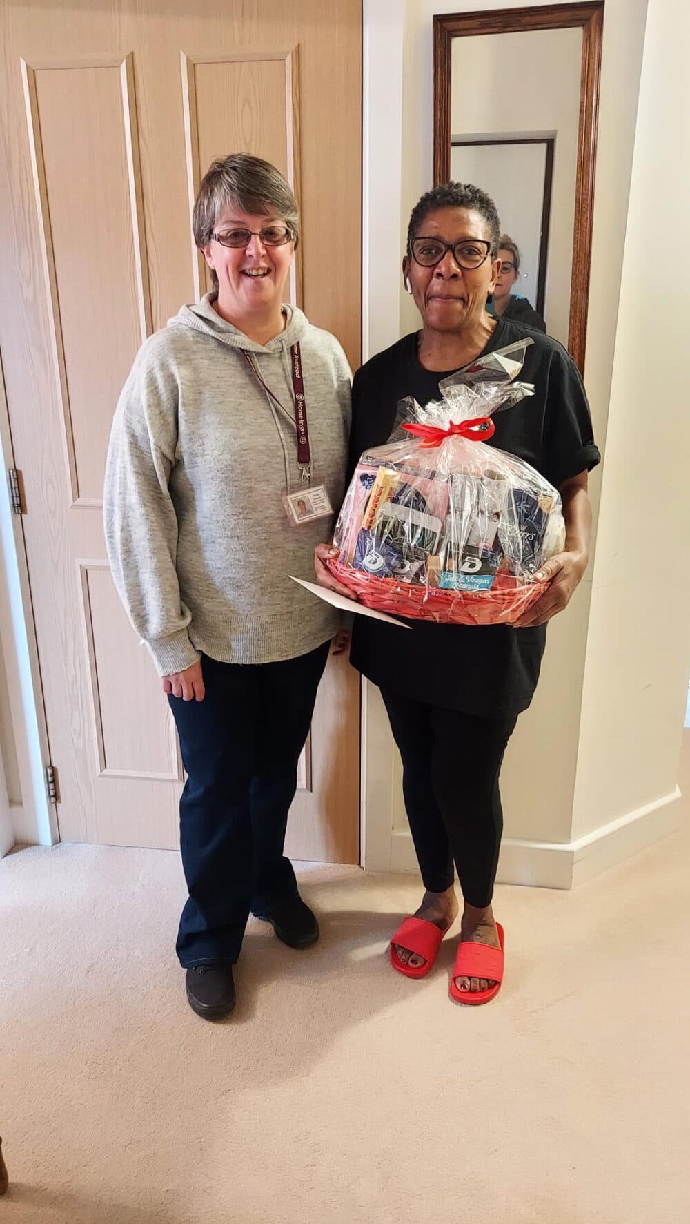Two women stand indoors, one holding a gift basket wrapped in cellophane and tied with a red ribbon. - Home Instead