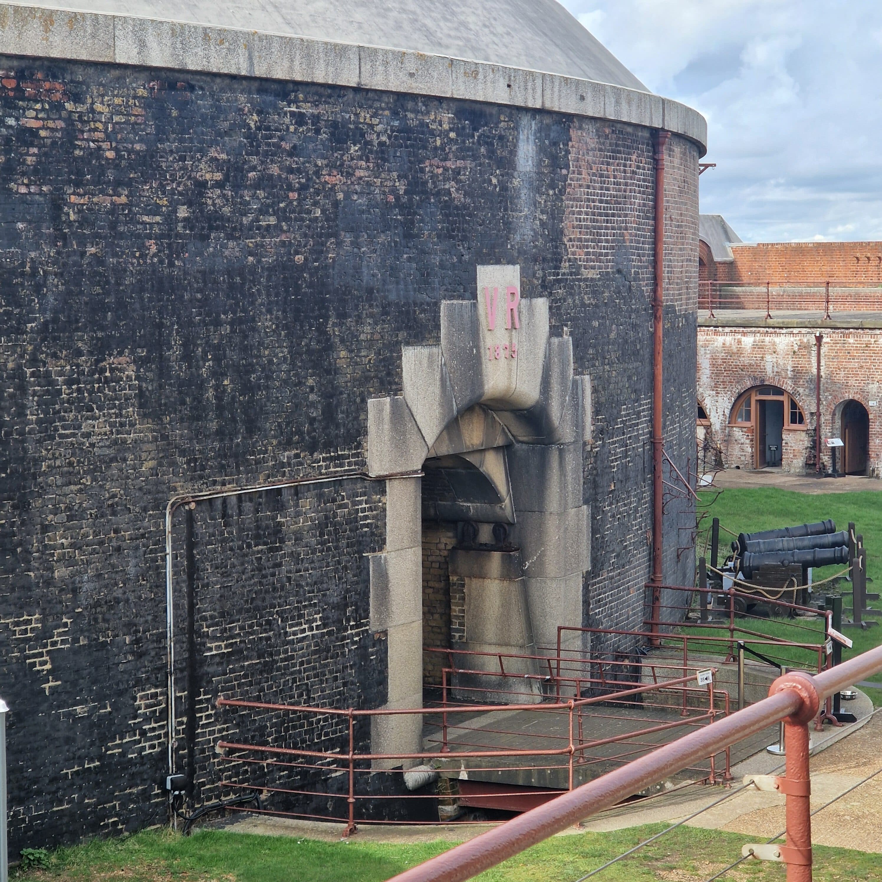 Large arched doorway in a round brick fort wall, with railings and a cannon on grass nearby. - Home Instead