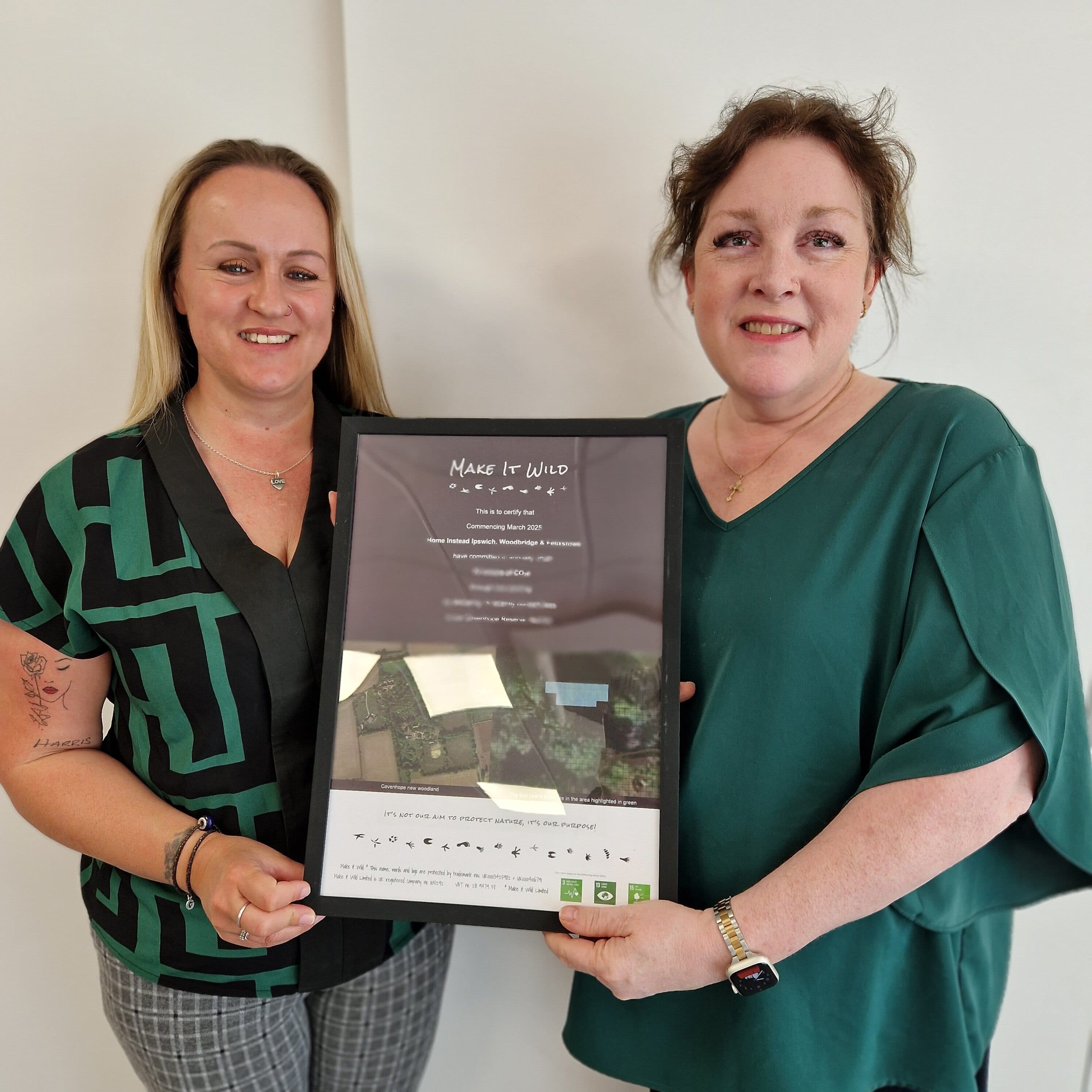 Two smiling women holding a framed "Make It Wild" certificate against a plain background. - Home Instead