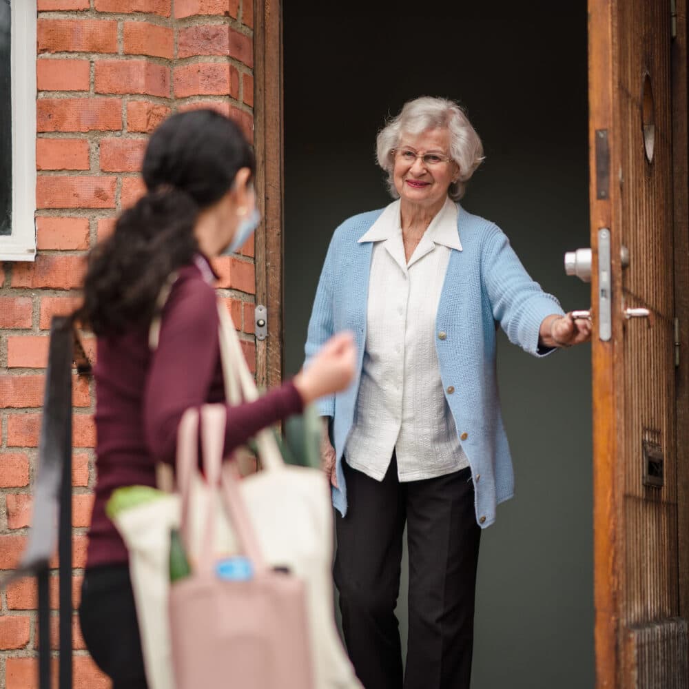 Home Instead client opening her front door for her Home Instead carer, who is arriving for a visit carrying shopping