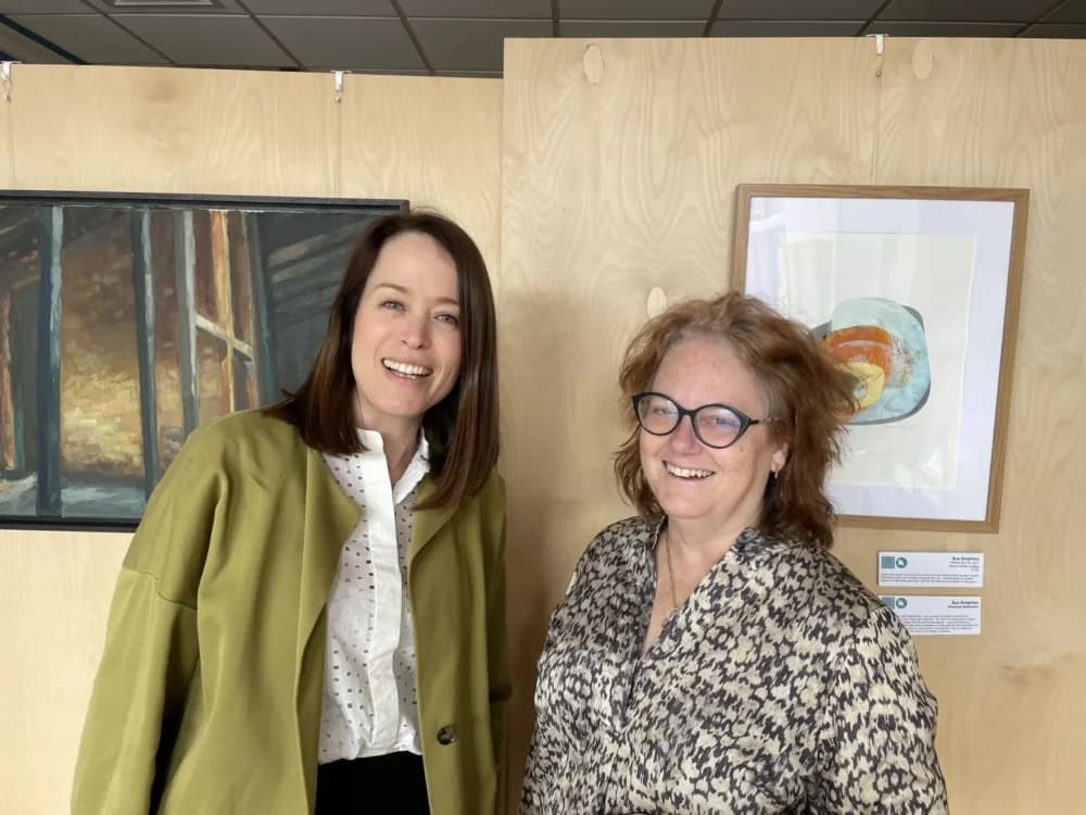 Two women smiling and standing in front of artwork displayed on wooden panels in a gallery setting. - Home Instead