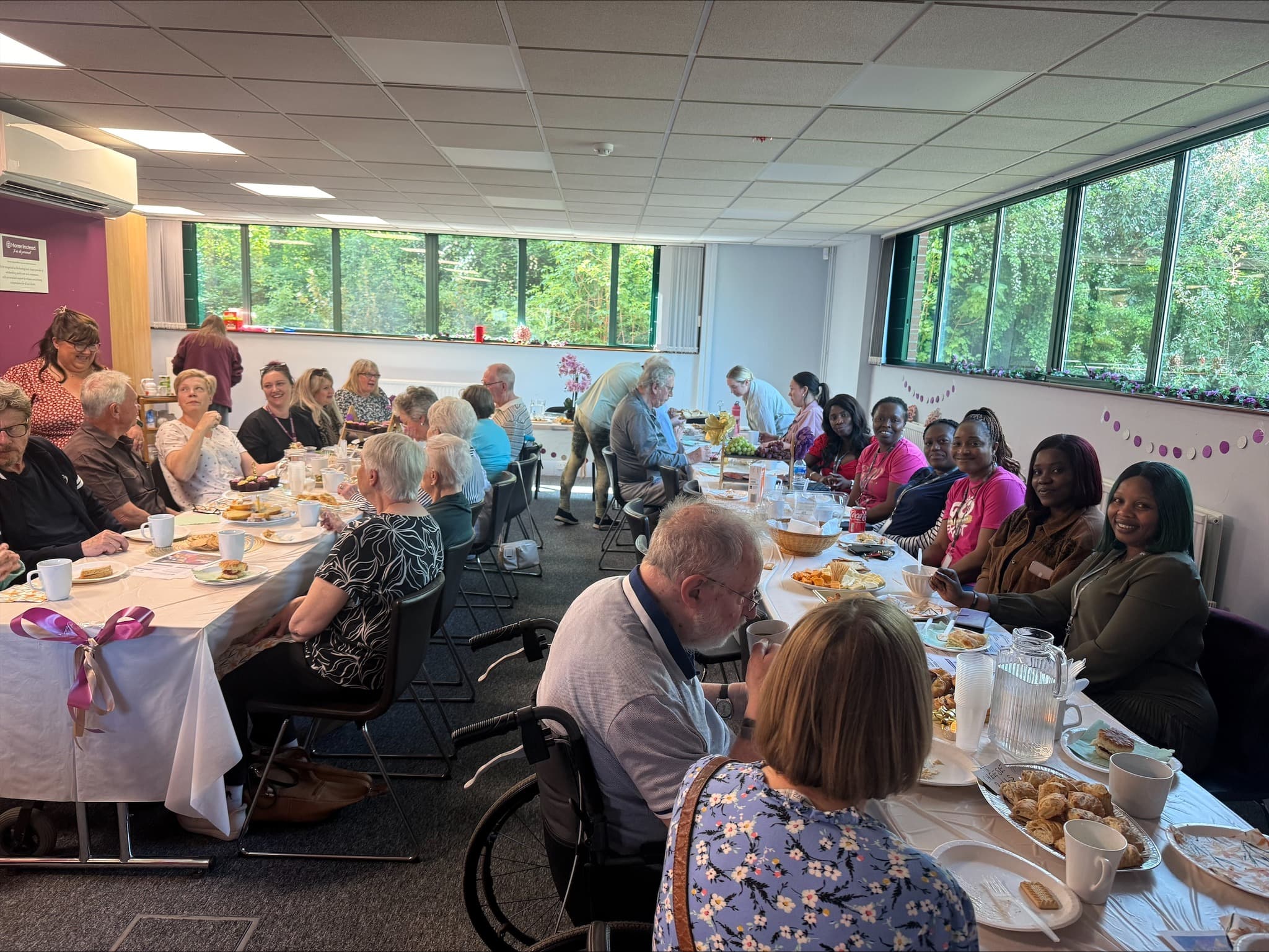 A group of people of all ages share food and conversation around long tables in a bright indoor room. - Home Instead