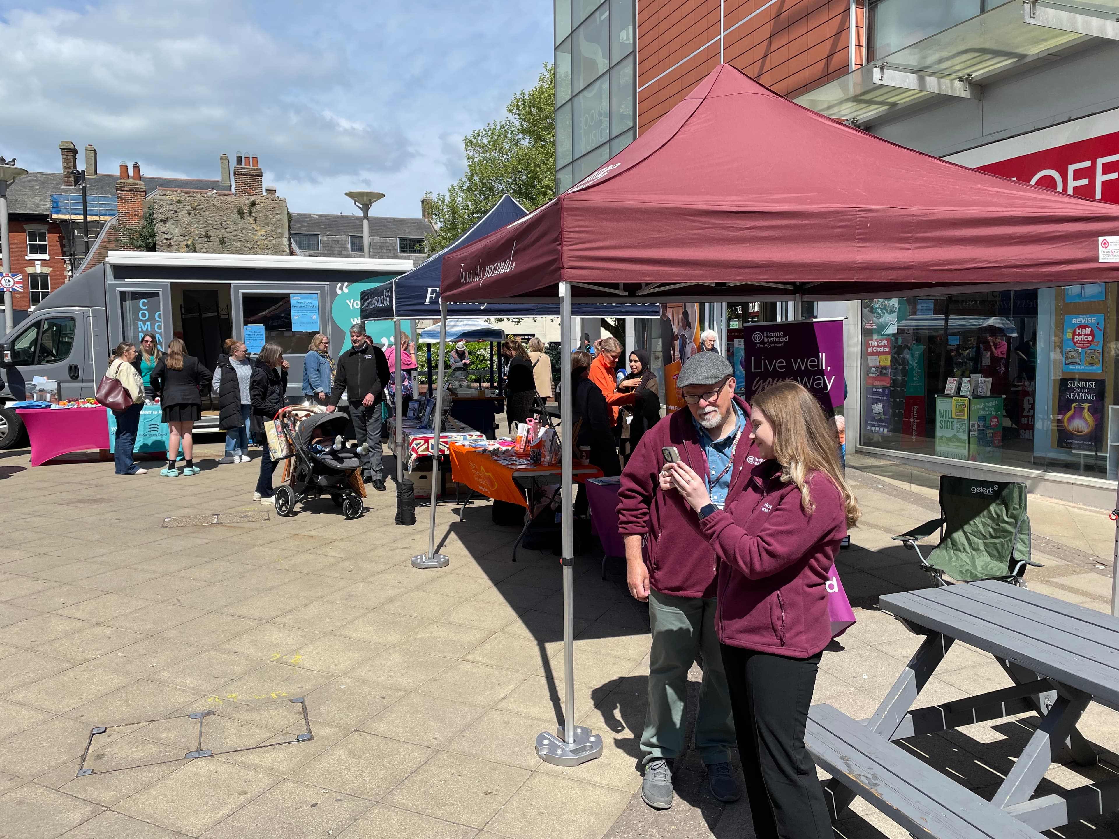People gather at outdoor market stalls under canopies on a sunny day near a modern building. - Home Instead