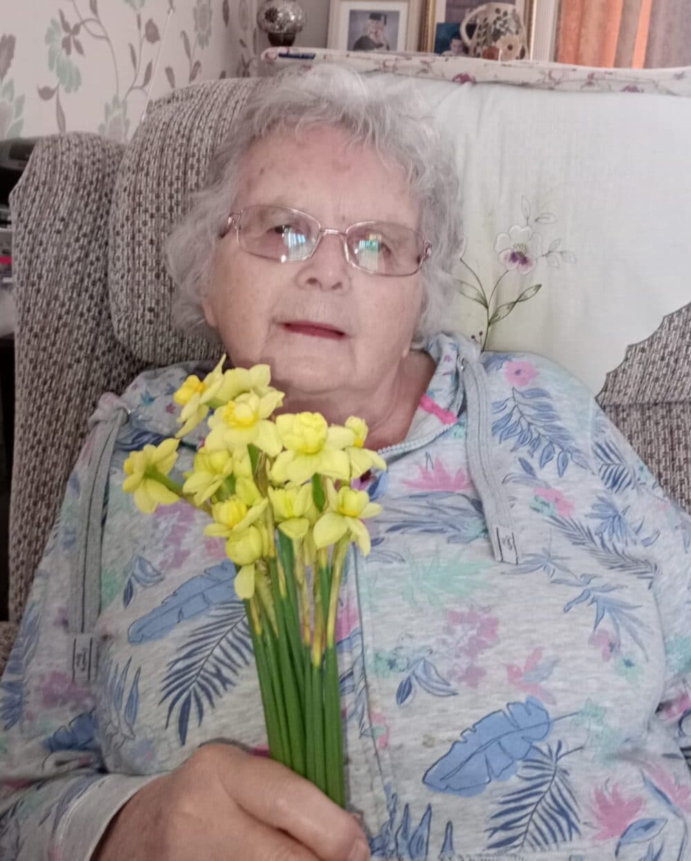 Elderly woman with grey hair holding yellow flowers, sitting on a patterned chair and smiling. - Home Instead