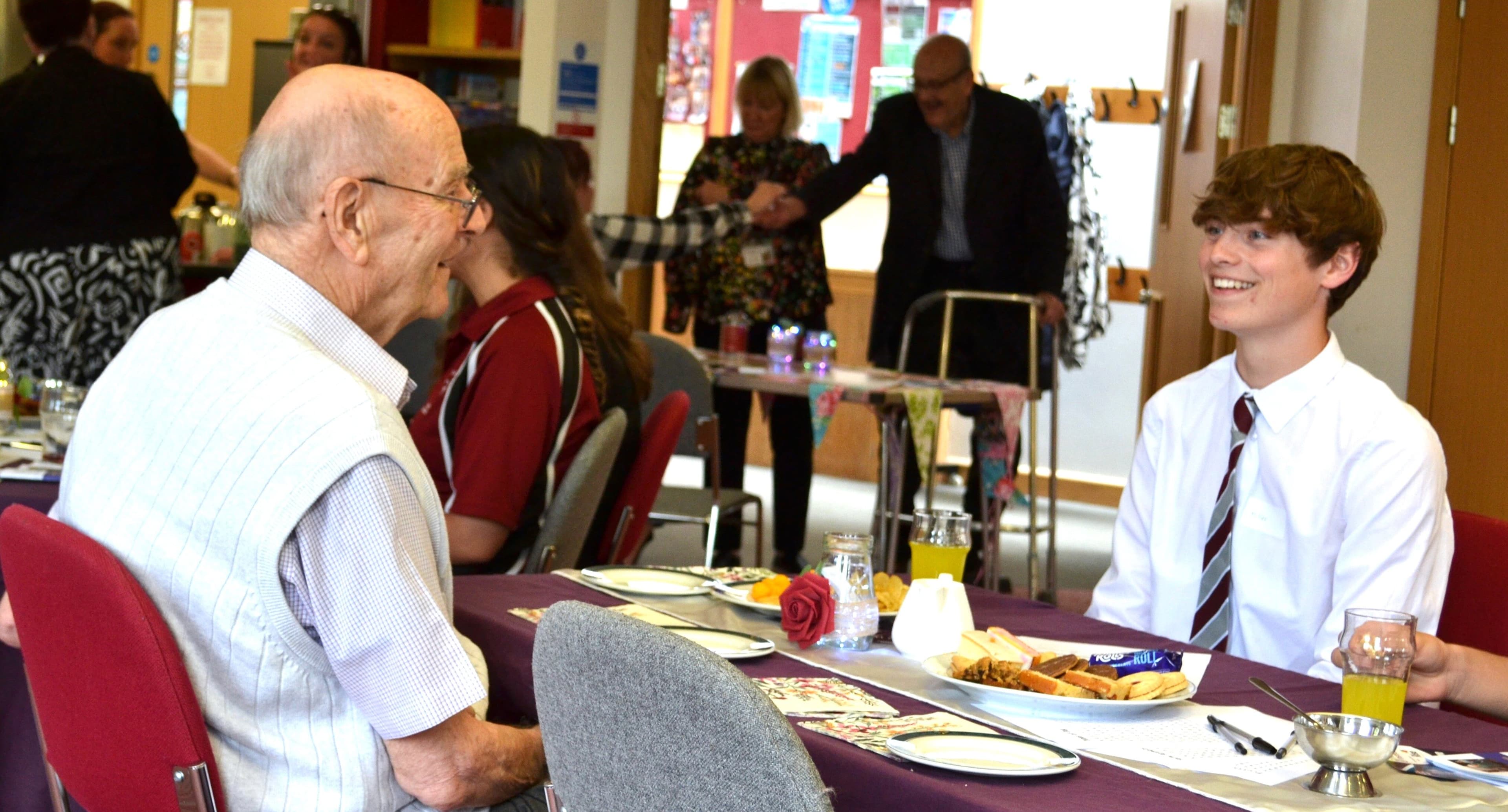 An elderly man and a teenage boy smile and talk at a table set for a meal in a community room. - Home Instead