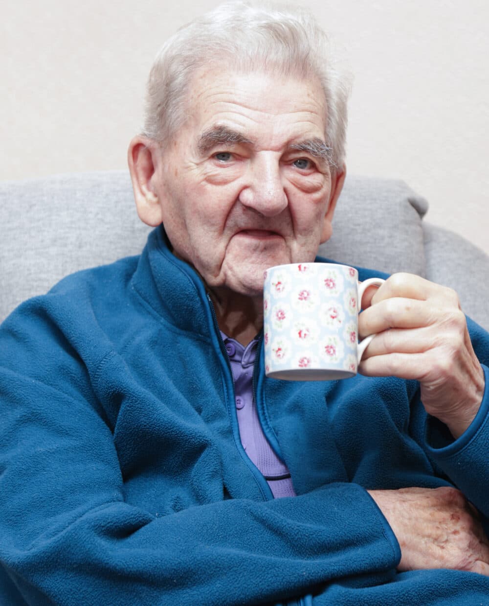Elderly man in a blue jacket sitting on a couch, holding a floral mug and smiling. - Home Instead