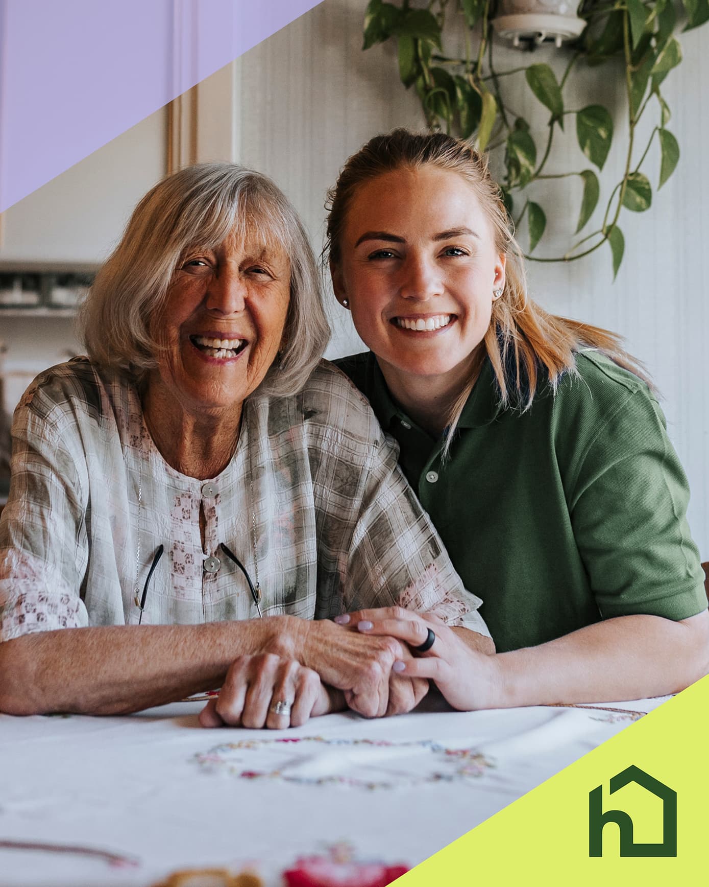 Smiling older woman and younger woman sit together at a table, holding hands, with a house logo in the corner. - Home Instead