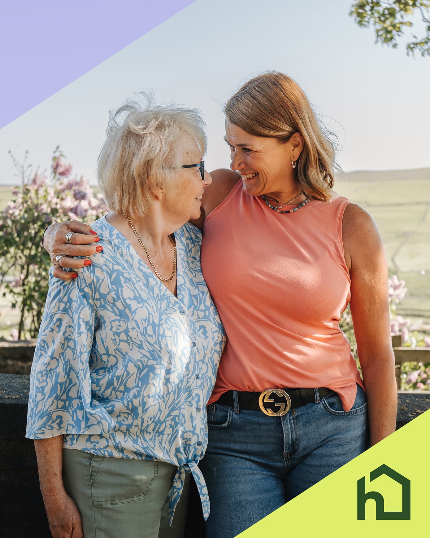 Two women stand outdoors, smiling and embracing each other, with a countryside background and a green house icon. - Home Instead