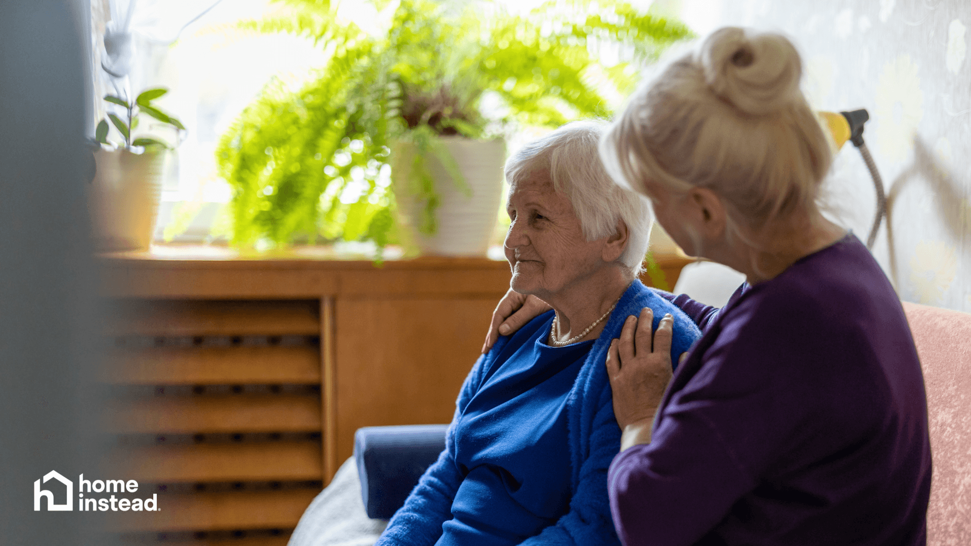 An elderly woman sits on a couch while another woman gently comforts her; homeinstead logo in corner. - Home Instead