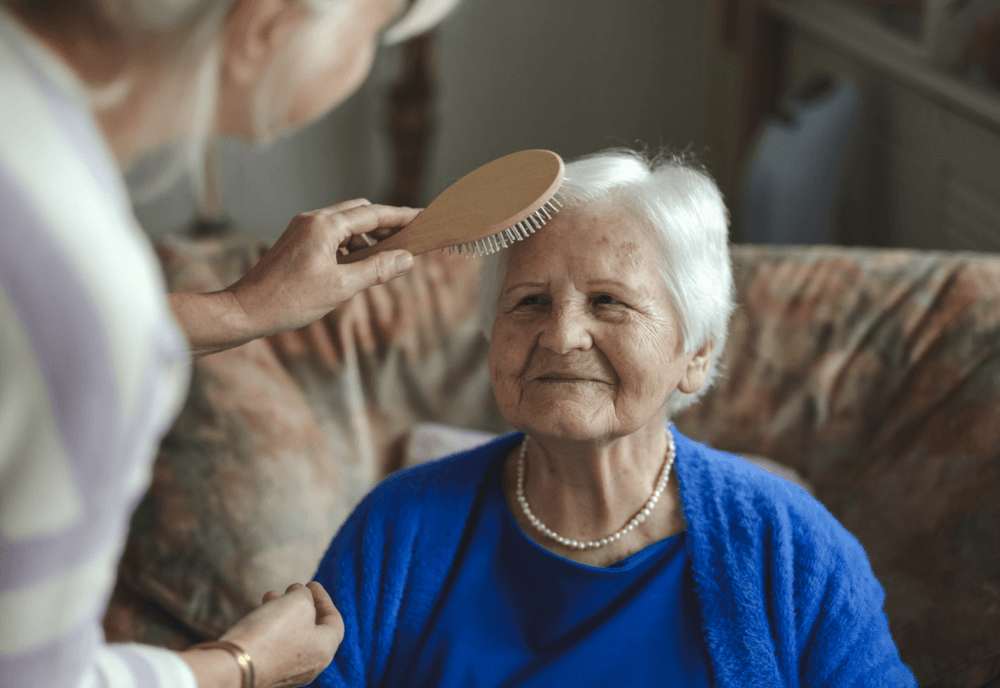 A woman brushes an elderly woman's hair as she sits on a sofa, smiling and wearing a blue cardigan. - Home Instead