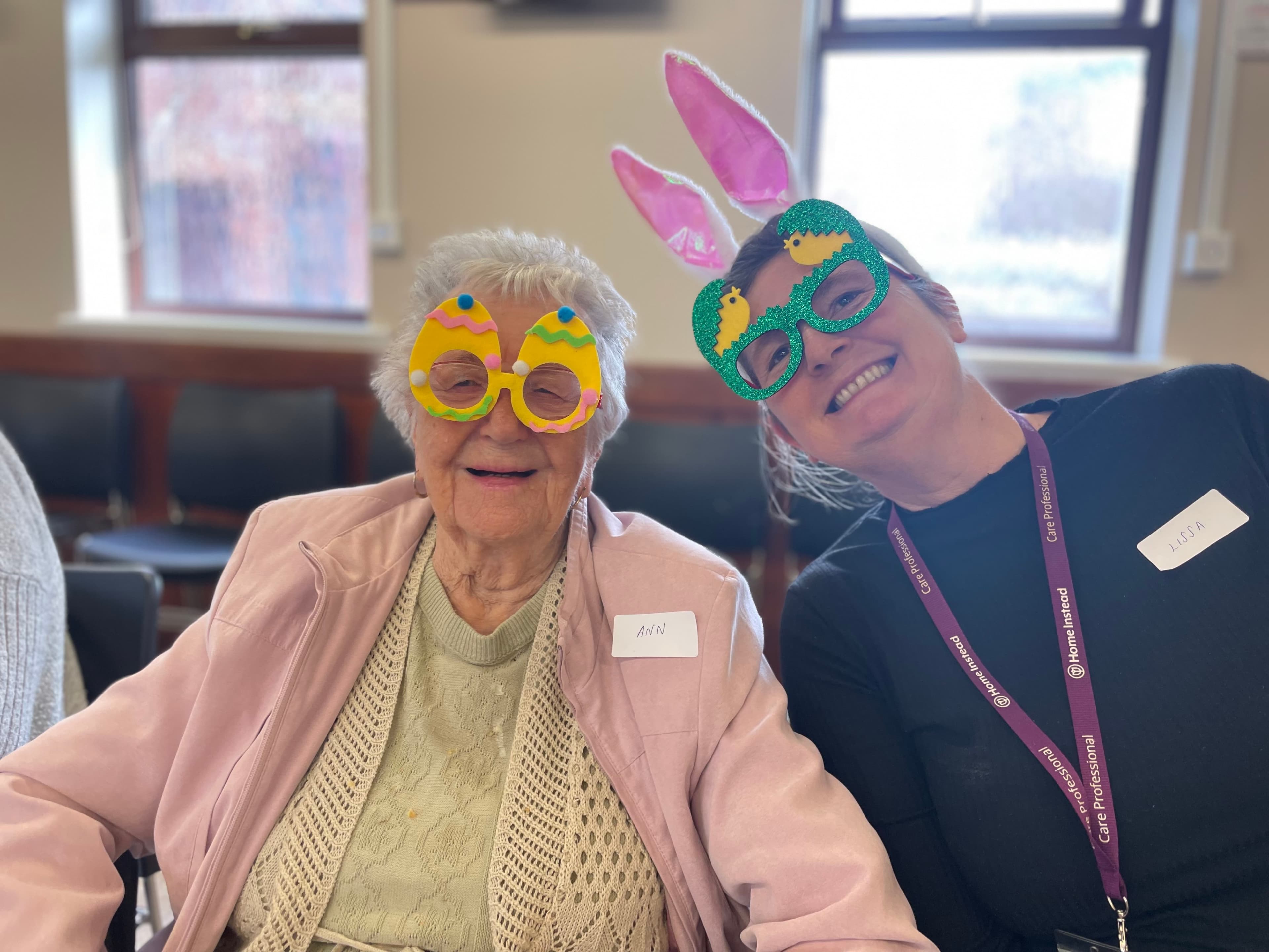 This image shows two people wearing festive Easter-themed novelty glasses at what appears to be a community event. On the left is an elderly woman with white hair wearing a pink jacket over a light yellow sweater and a name tag that reads "ANN." She's smiling and wearing yellow egg-shaped glasses with colorful decorations. On the right is a care professional wearing a dark uniform with a Home Instead lanyard and name tag, sporting green sparkly glasses with pink bunny ears. They're sitting in what looks like a community hall with windows visible in the background. The photo captures a joyful moment of connection during what seems to be an Easter celebration activity.