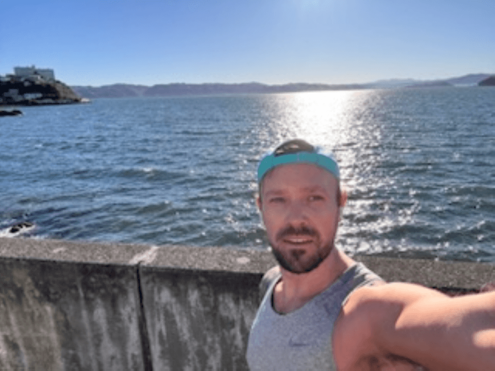 images shows a man taking a selfie against a blue water background. Clear sunny sky with mountains in the distance.