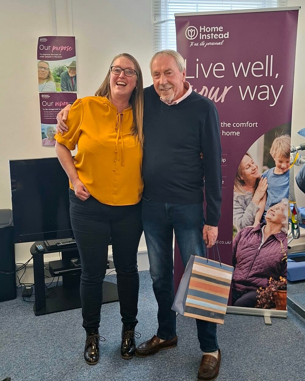 Two people smiling and posing together indoors, with a gift bag and "Home Instead" banner behind them. - Home Instead