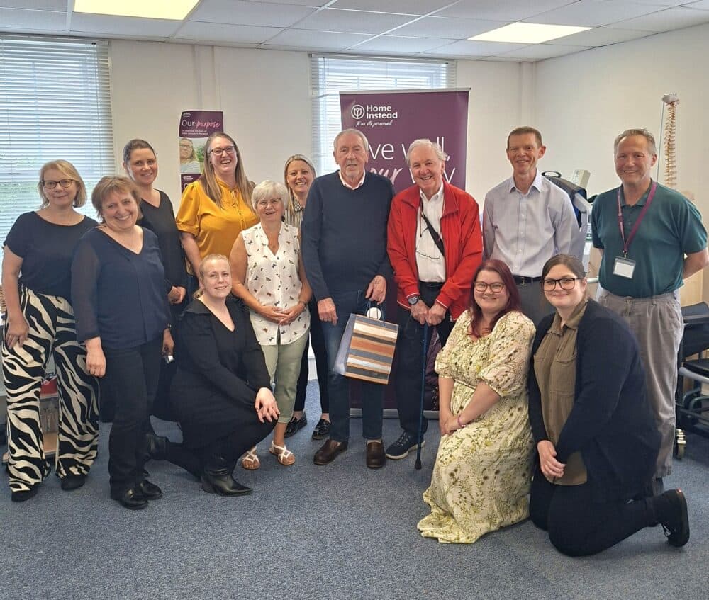 A group of people smiling and posing together in an office-style room with banners in the background. - Home Instead