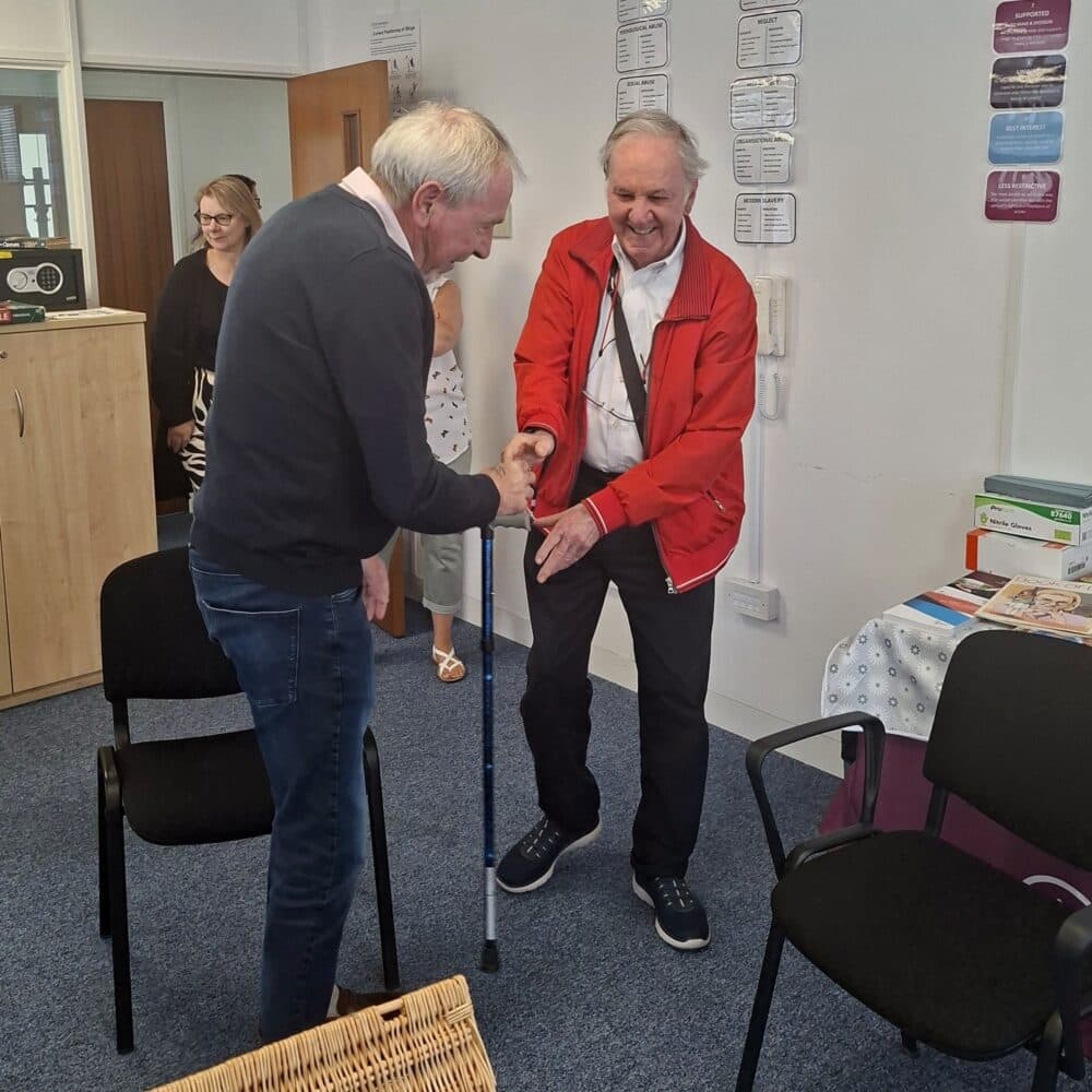 Two older men shake hands and smile in a room, with two women and empty chairs in the background. - Home Instead