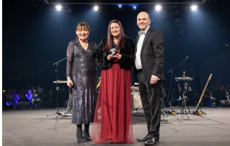 Three people in formal attire stand on stage at an award ceremony, smiling at the camera. - Home Instead