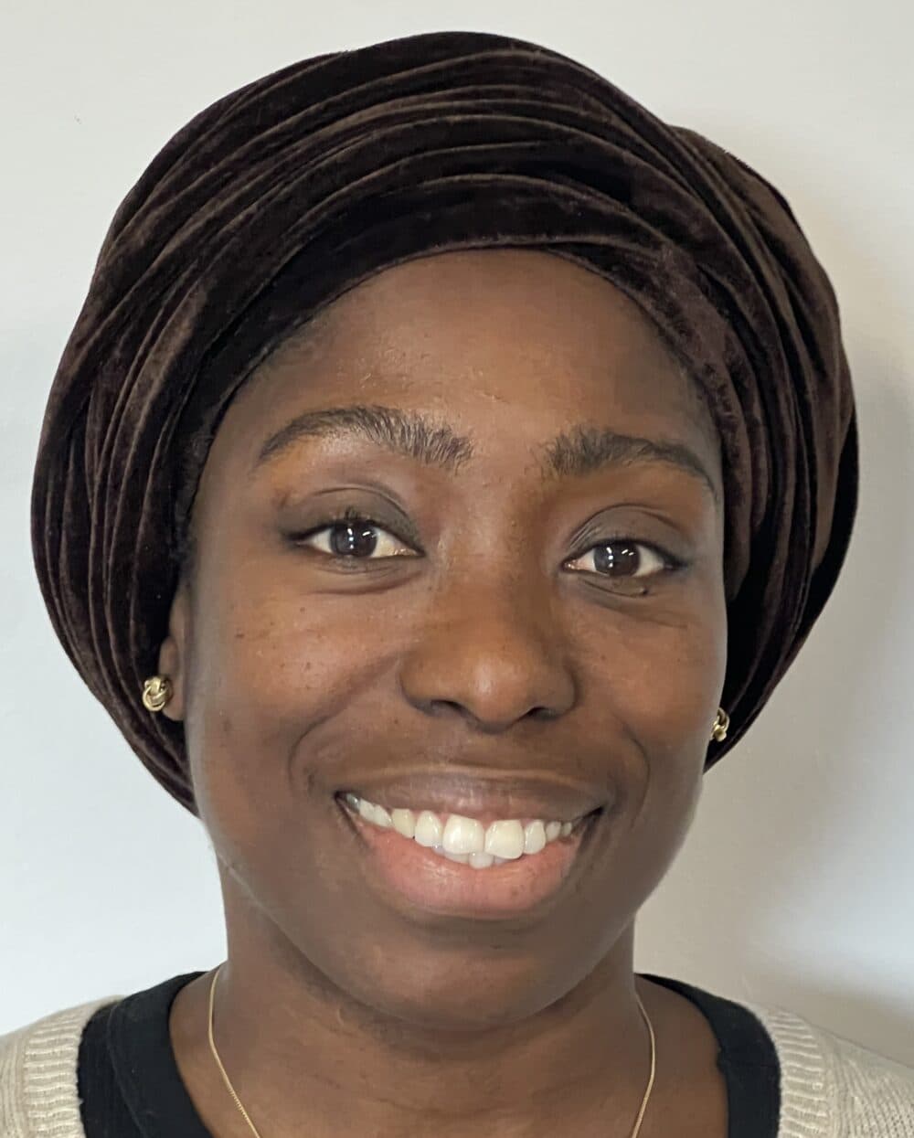 Woman smiling, wearing a dark velvet headwrap, light sweater, and gold earrings, against a plain background. - Home Instead