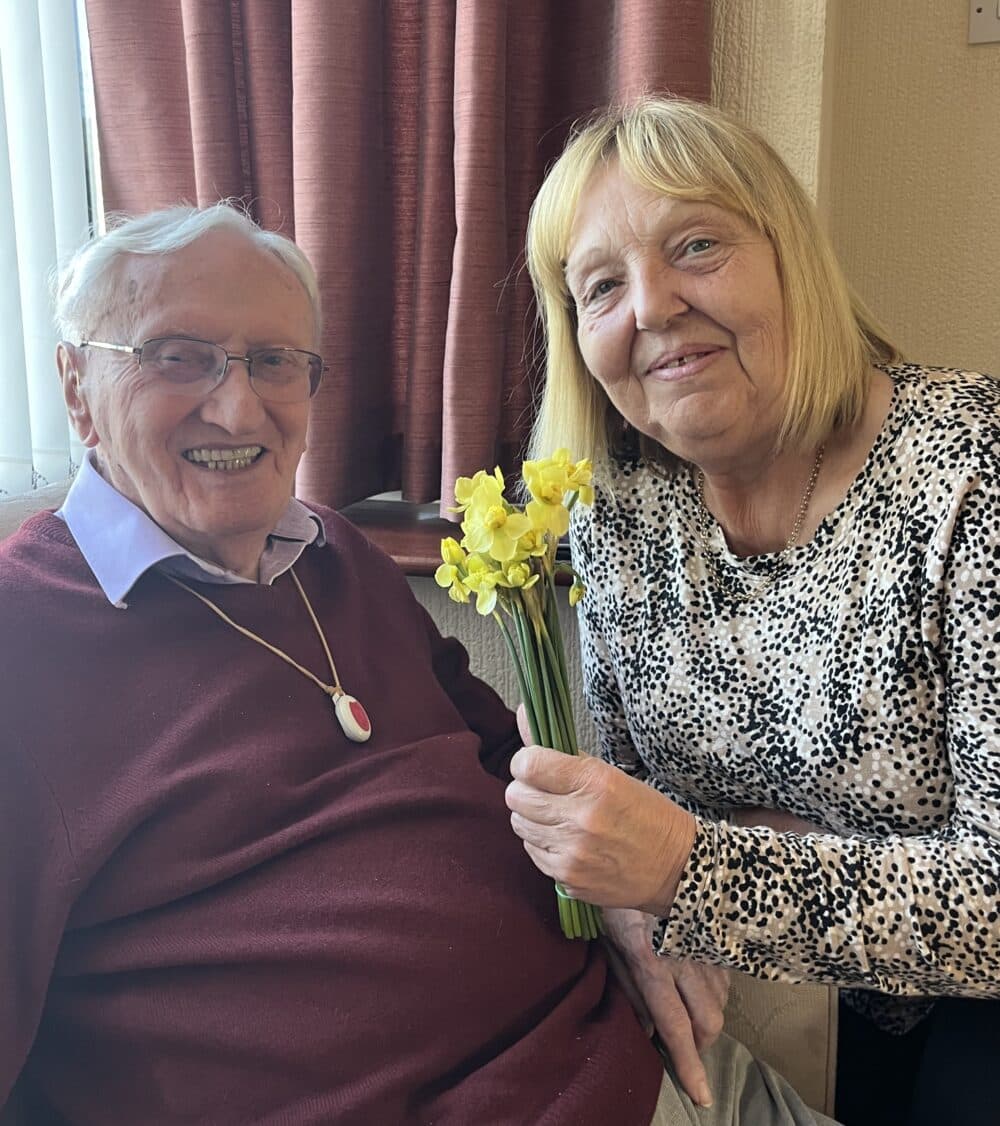 Elderly man and woman smiling together; woman holds a small bunch of yellow daffodils. - Home Instead