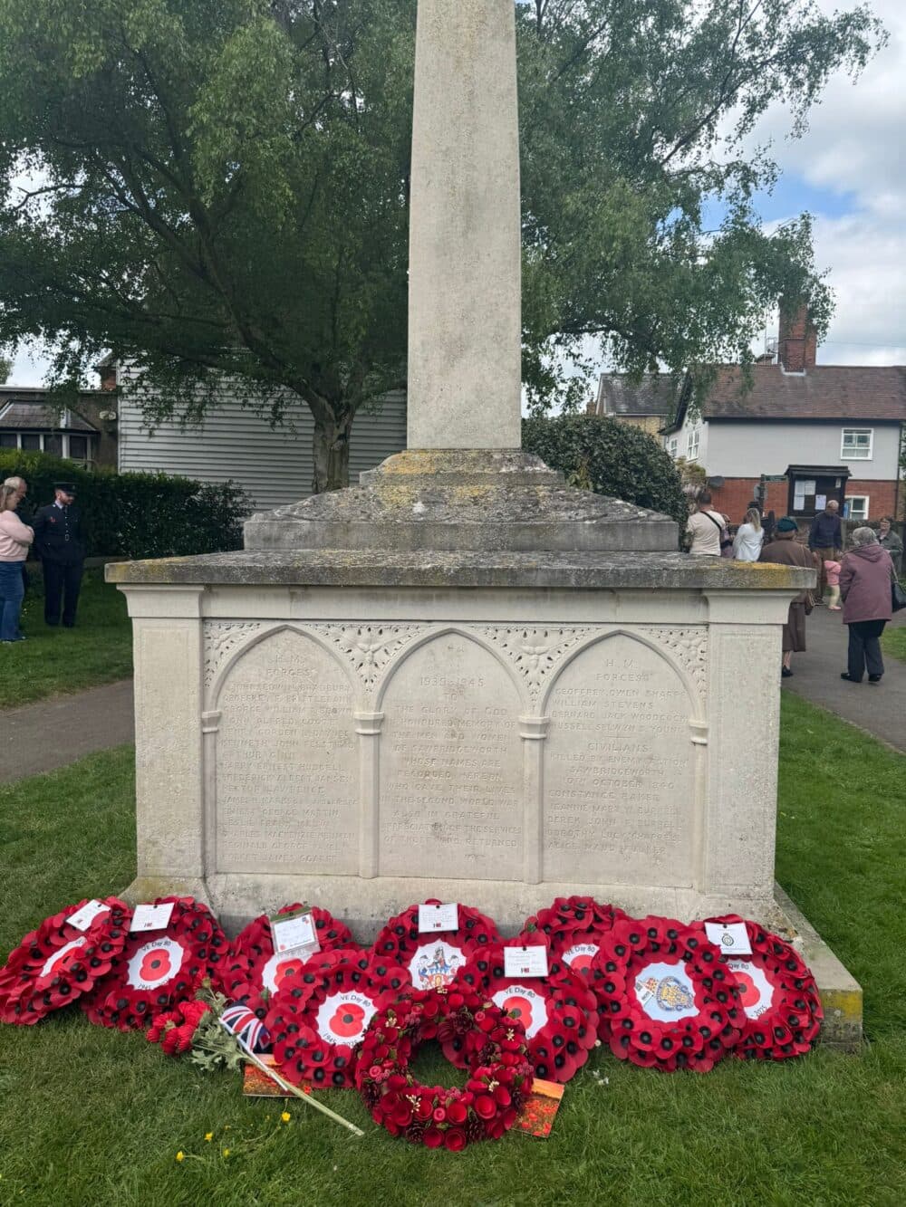A stone war memorial with red poppy wreaths at its base, surrounded by people and trees. - Home Instead