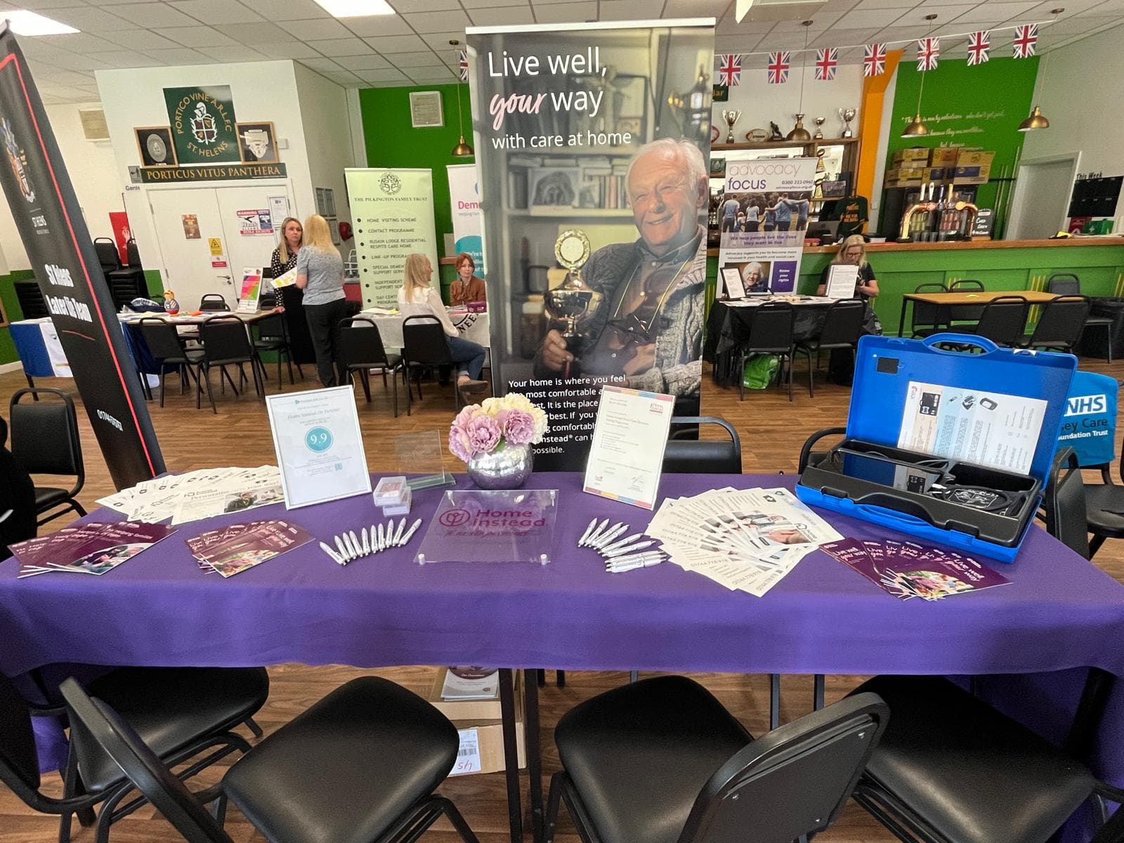 Image shows a purple desk with pens, adverts and awards on. This is set in a hall with other displays in the background.