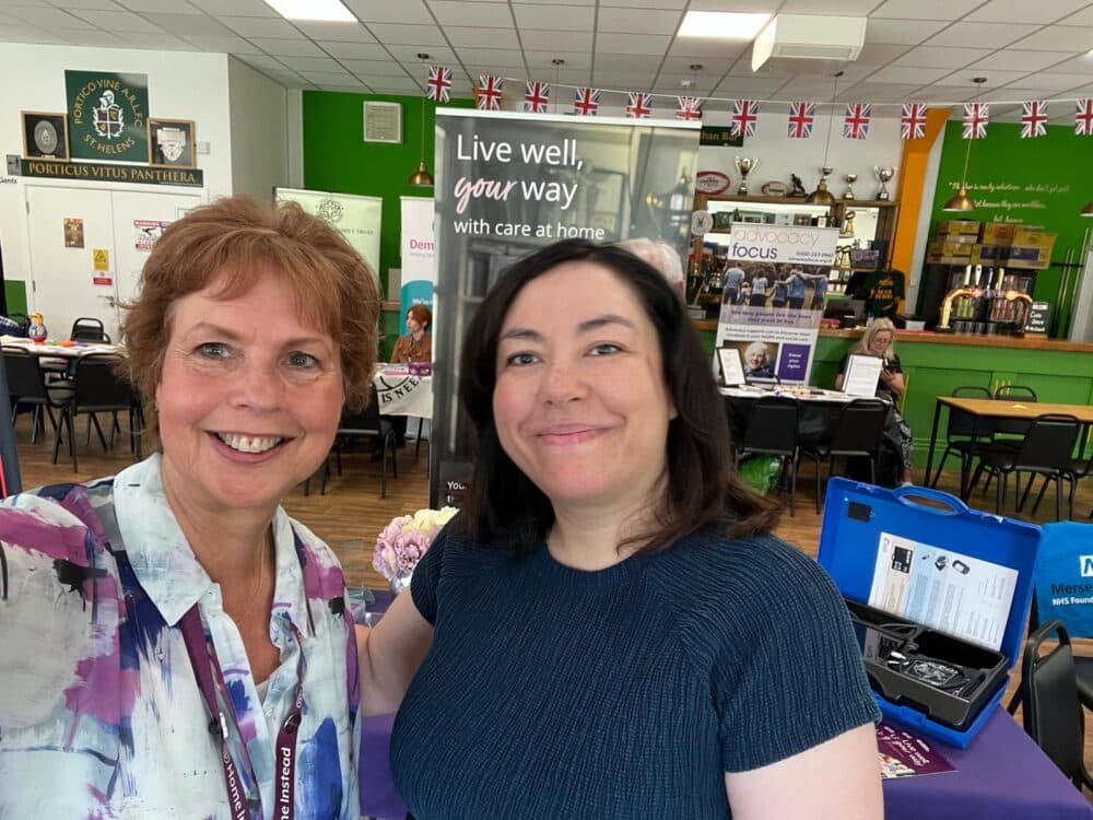 Image shows 2 ladies taking a picture. One is in a colourful blouse and the other is in a dark top. They are in a hall with displays behind them.