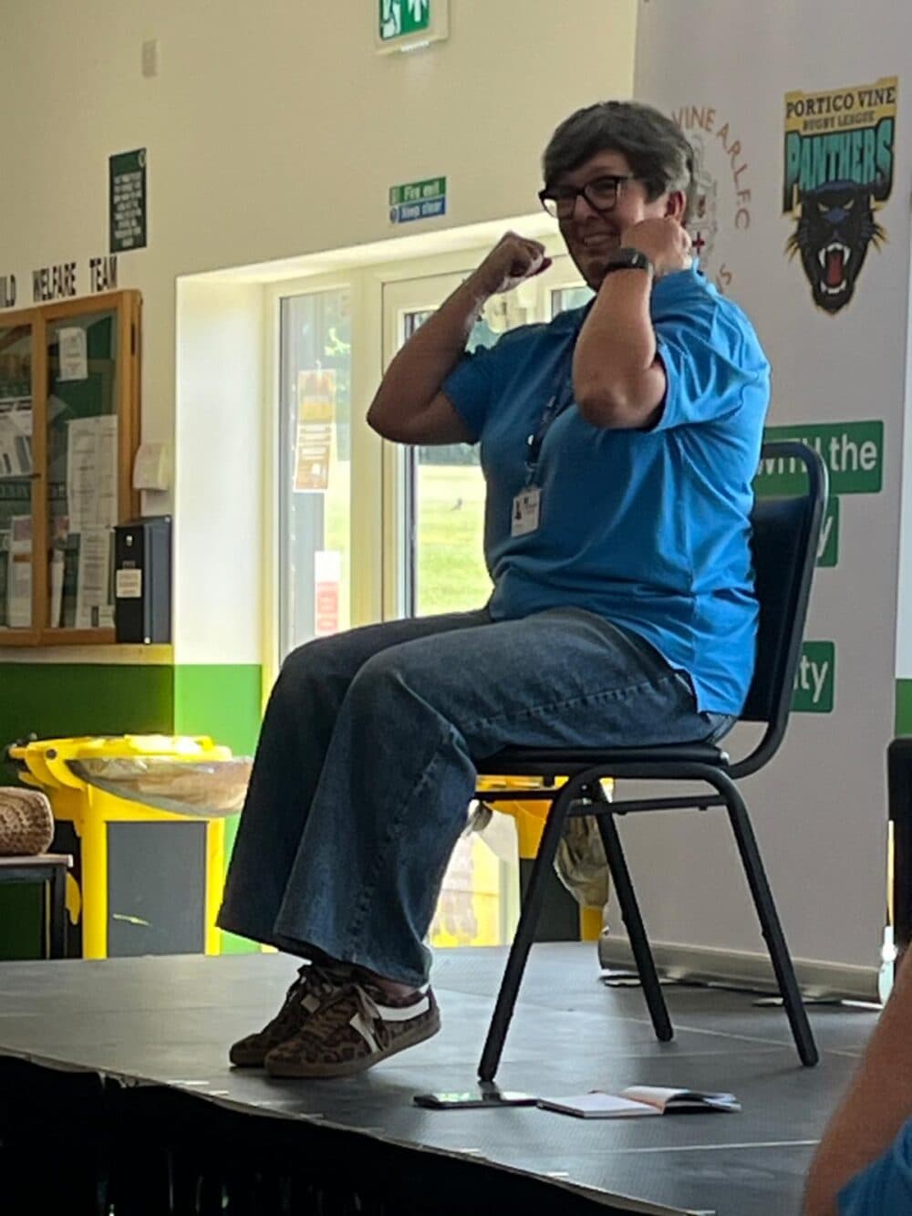 Image shows a lady sat on a chair demonstrating chair exercises. She is on stage.