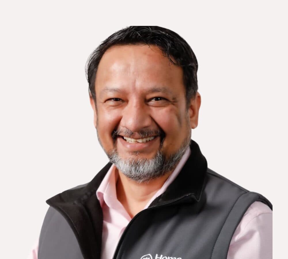Smiling man with short dark hair, beard, and a grey vest over a light pink shirt on a plain background. - Home Instead
