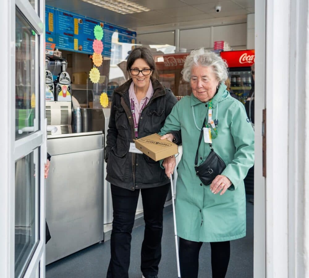 An older female adult with white hair wearing green coat going out of the store and using a crane with her younger female carer wearing eyeglasses and with long hair