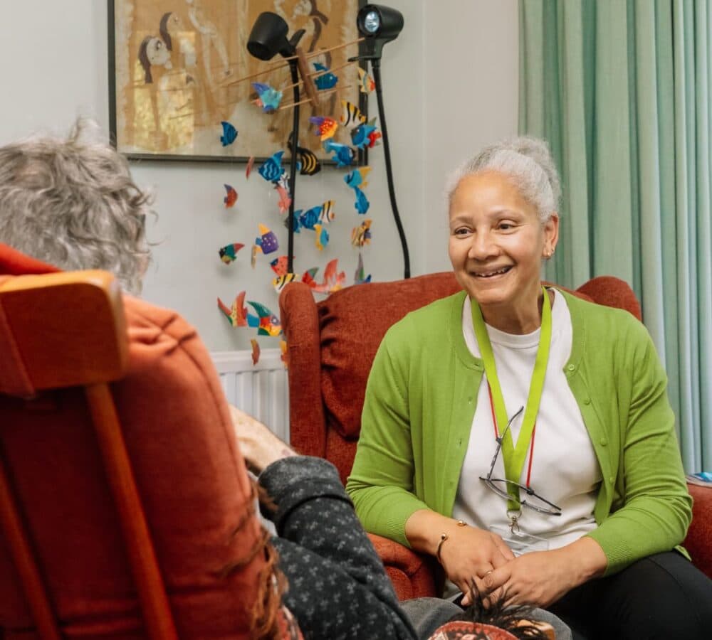 An older female adult wearing green and with white hair smiling and chatting with an older adult while sitting on a couch inside the house