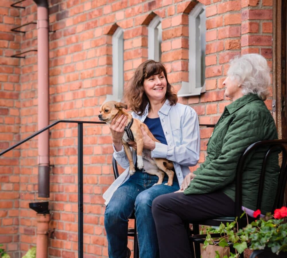 Two women chatting outside the house both happy and smiling while the younger woman is holding a dog too