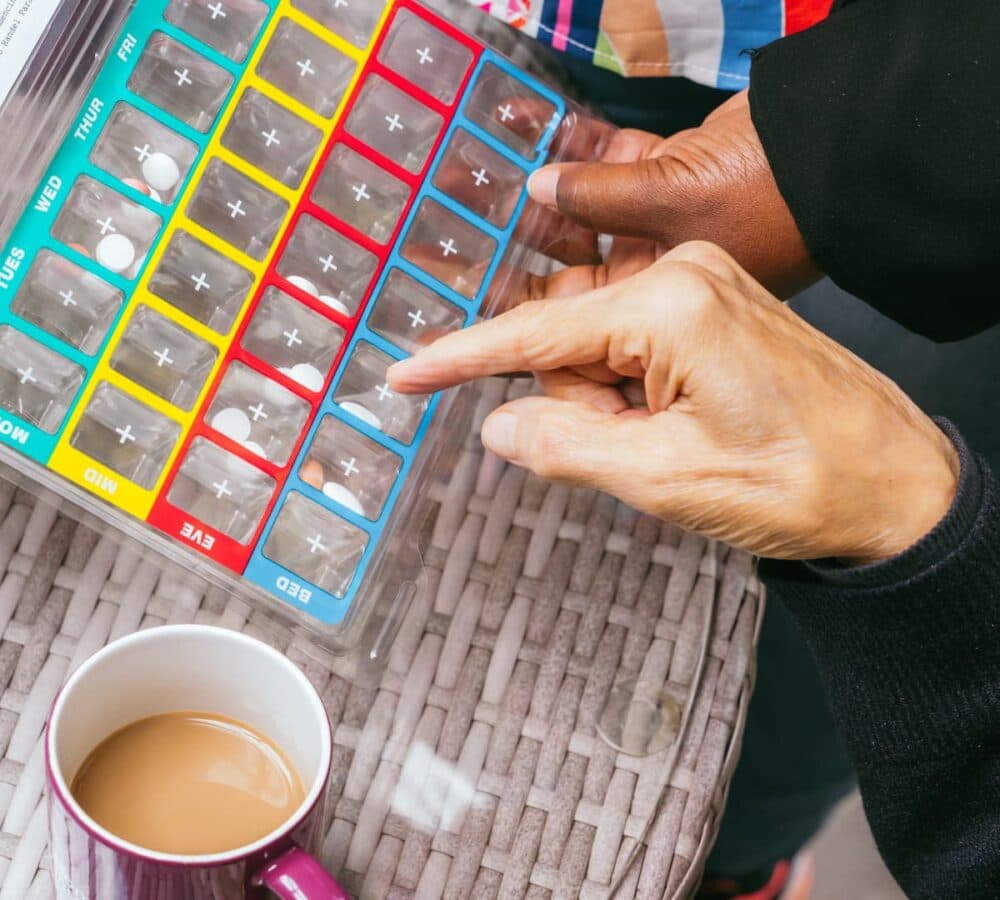 Medicine organiser in different colours with a cup of coffee on the side and two hands pointing