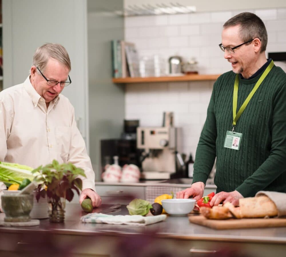 a male carer wearing green and chopping vegetables in the kitchen while chatting with a male older adult wearing eyeglasses and with grey hair both happy and smiling