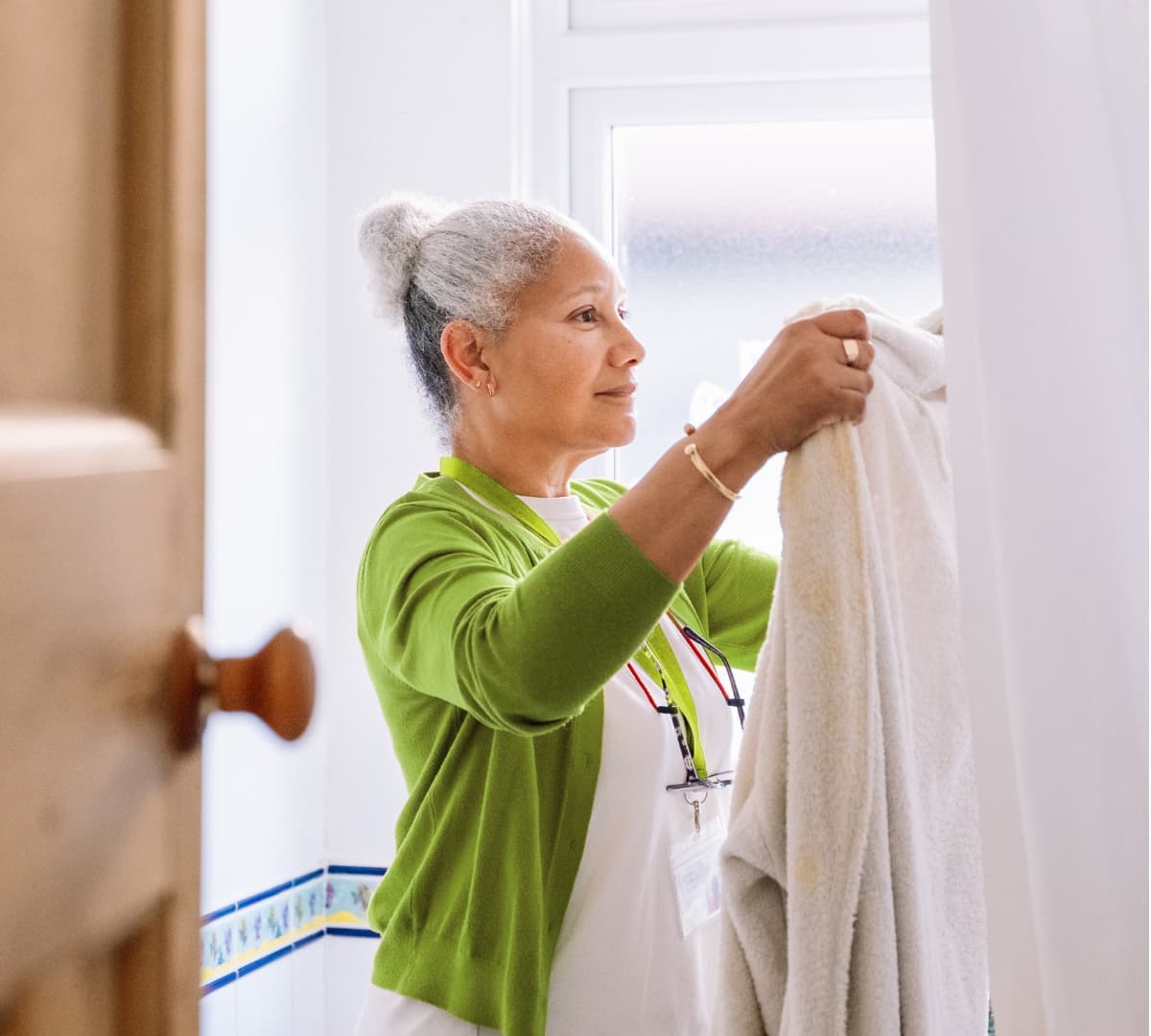 Older woman in a green cardigan holds a white towel in a bright, tiled bathroom. - Home Instead