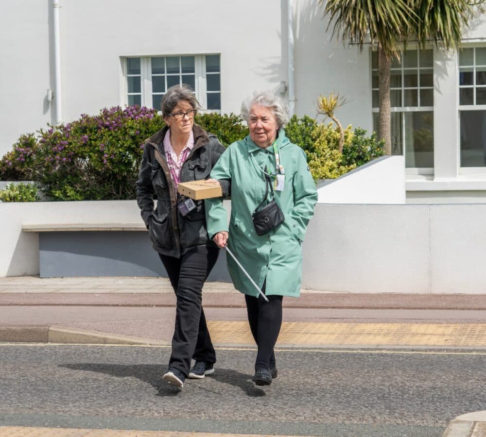 An older female adult with white hair wearing green coat crossing the road and using a crane with her younger female carer wearing eyeglasses and with long hair