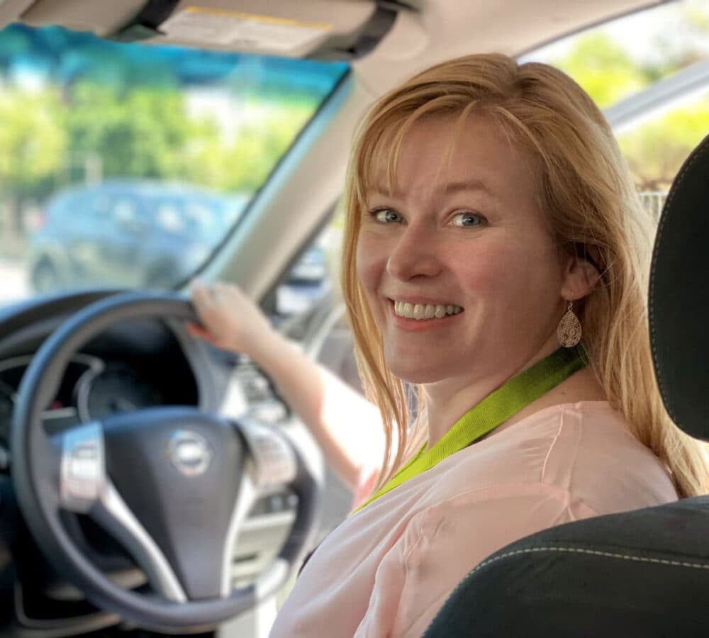 A young female with blonde hair and wearing pink smiling while driving a car