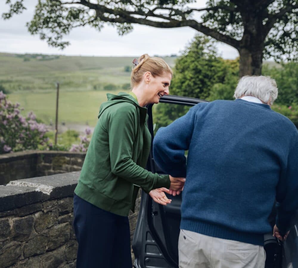 An older male adult with grey hair and wearing blue pullover sweater going inside the car with his younger female carer with blonde hair and wearing green swetashirt both going inside the car