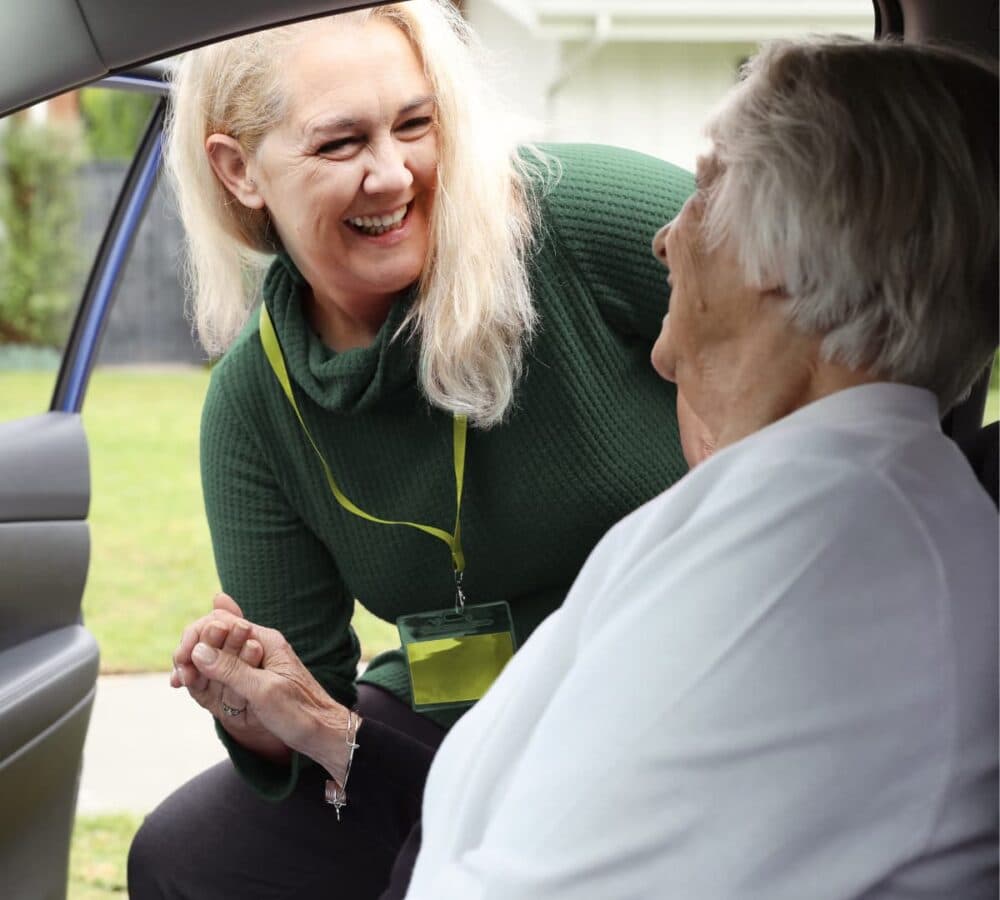 An older female adult wearing white getting out of the car with the help of her younger female carer