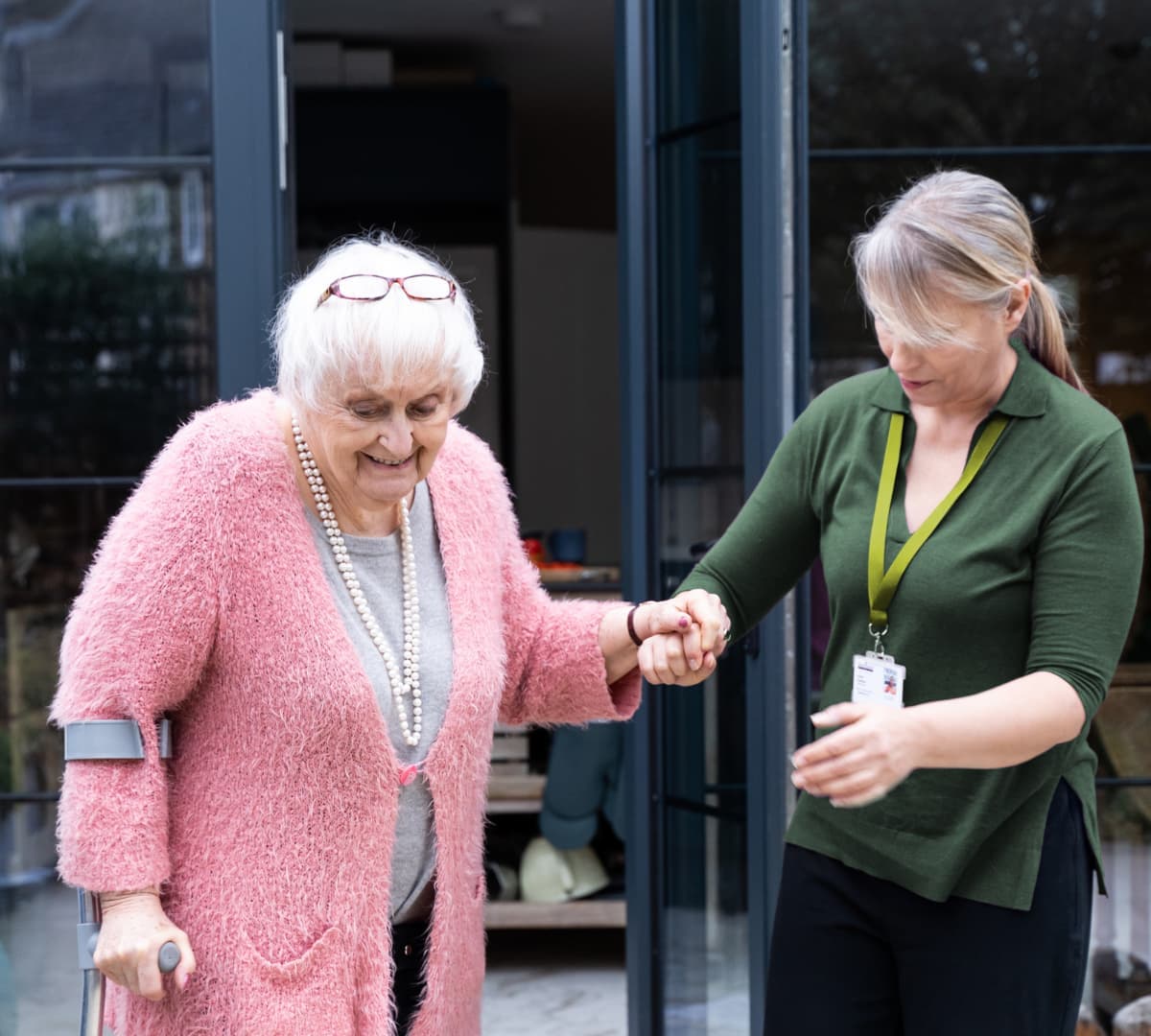 An older female adult wearing pink and with white hair and usung a crutch going out the door with the help of her younger female carer wearing green