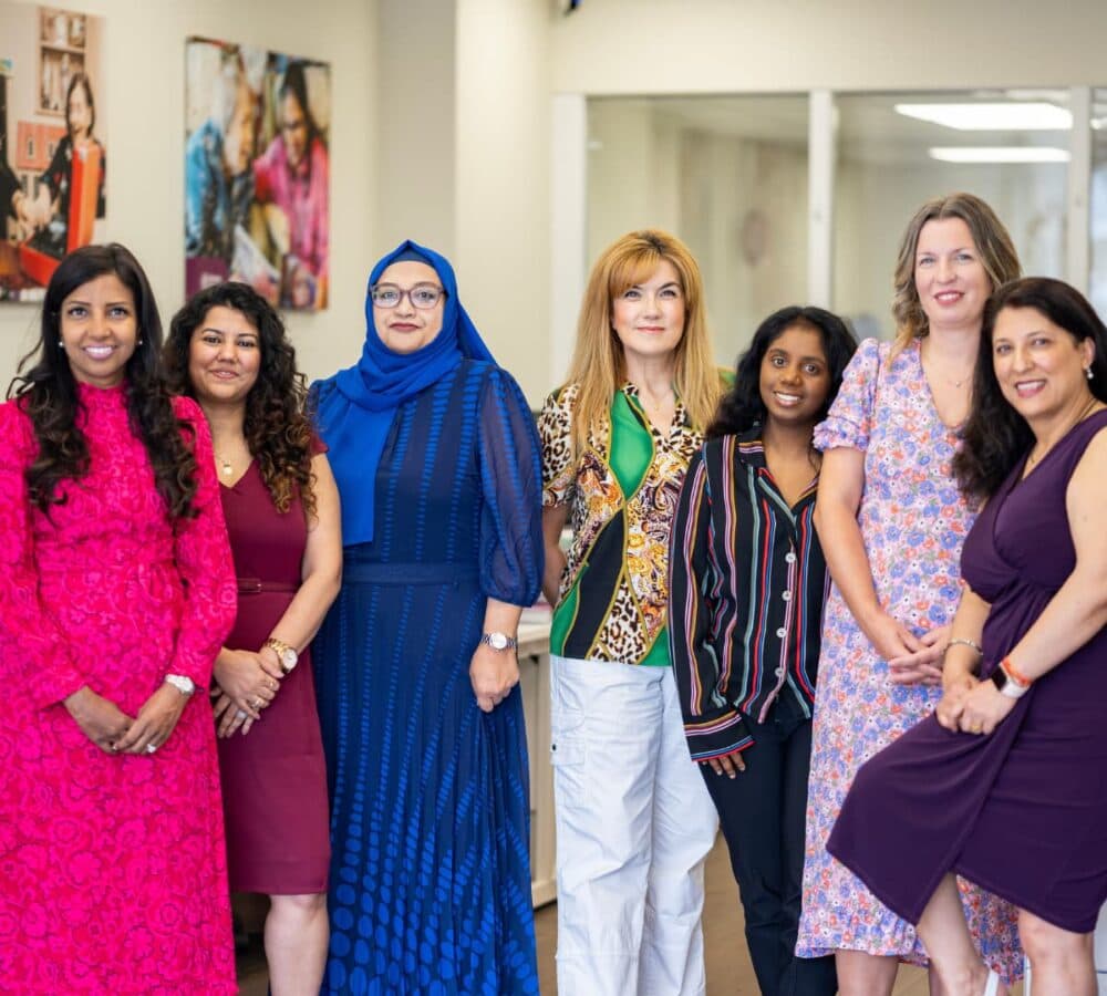Seven women standing side by side indoors, smiling, dressed in colorful and diverse outfits. - Home Instead