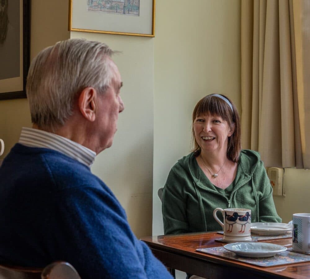 An older male adult sitting on a couch and having coffee in a green mug with his female carer with black hair and wearing skirt inside the house