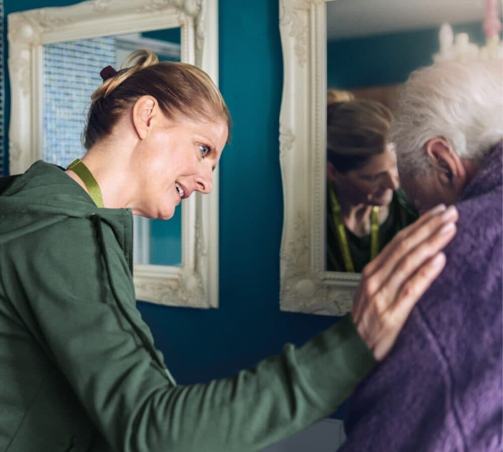 A female carer wearing green and smiling while helping an older male adult inside the bathroom