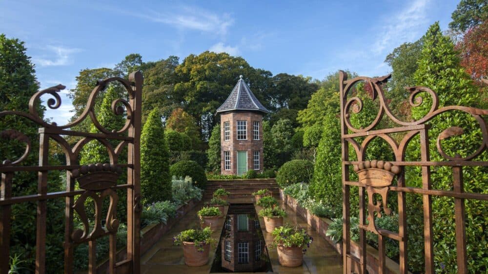 Ornate iron gates open to a formal garden with a brick turret and lush greenery under a blue sky. - Home Instead
