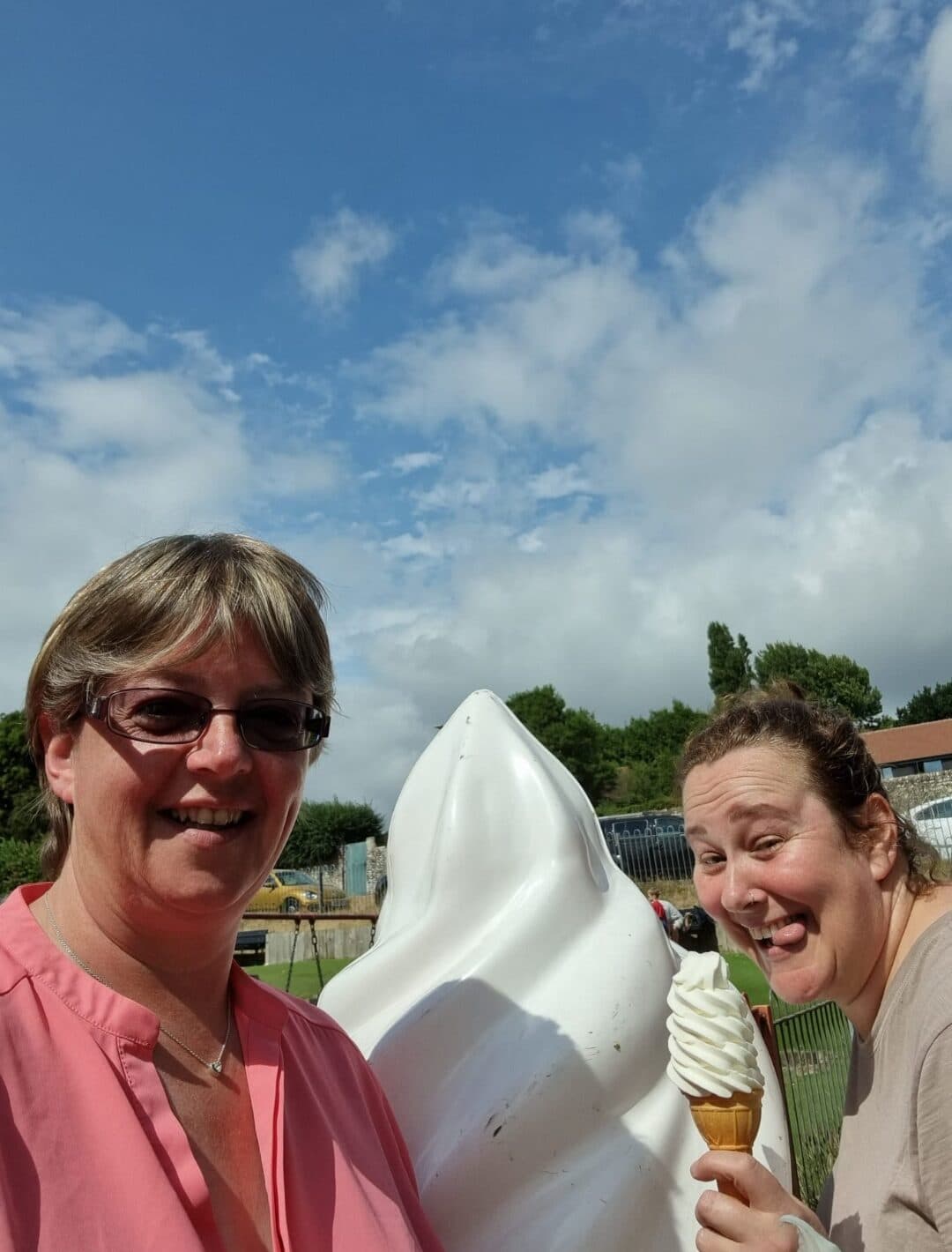 Two smiling women pose outdoors with a giant ice cream sculpture and a real ice cream cone under a blue sky. - Home Instead
