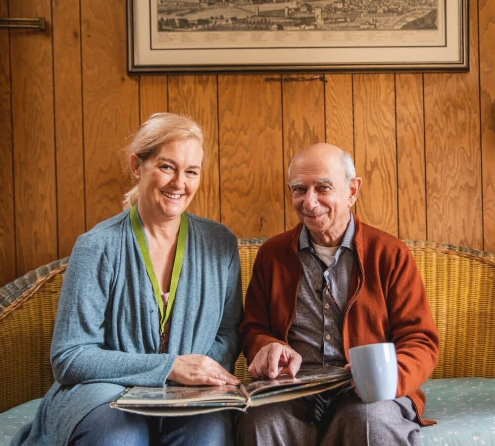An elderly man and woman sit on a couch, smiling and looking at a photo album together. - Home Instead