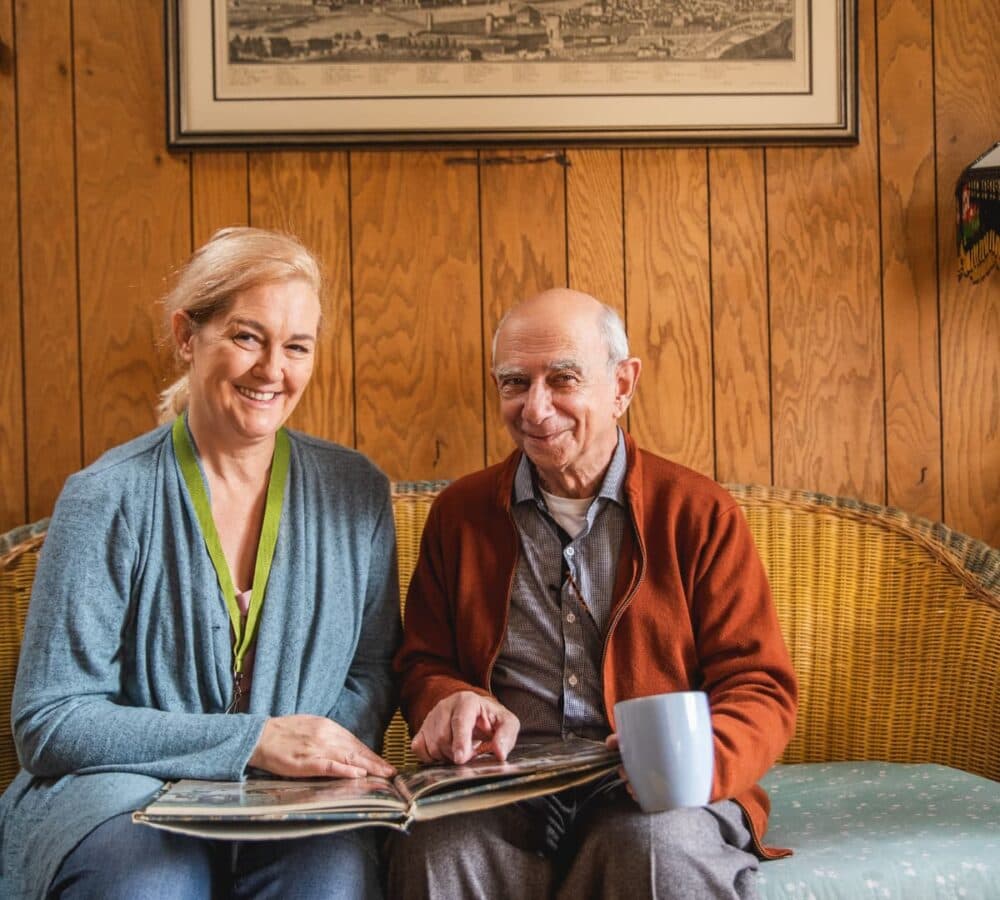 An older male adult with grey hair sitting on a couch with his younger female carer wearing blue and with blonde hair both happy and smiling while looking at a photo album
