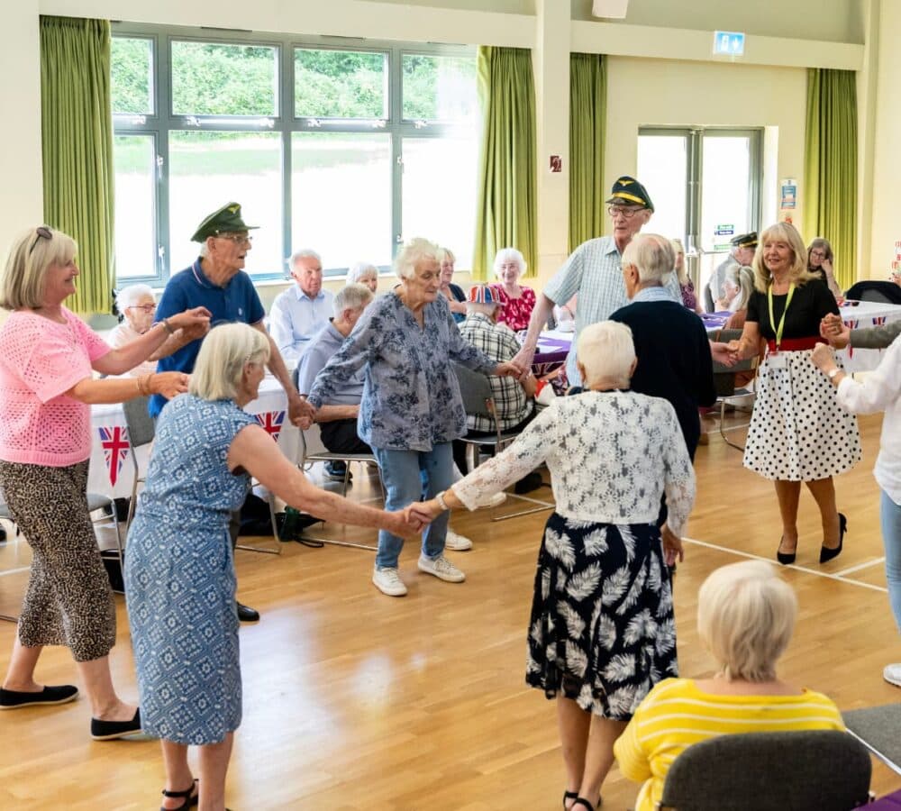 A group of older adults holding hands and dancing in a bright community hall decorated with Union Jack flags. - Home Instead